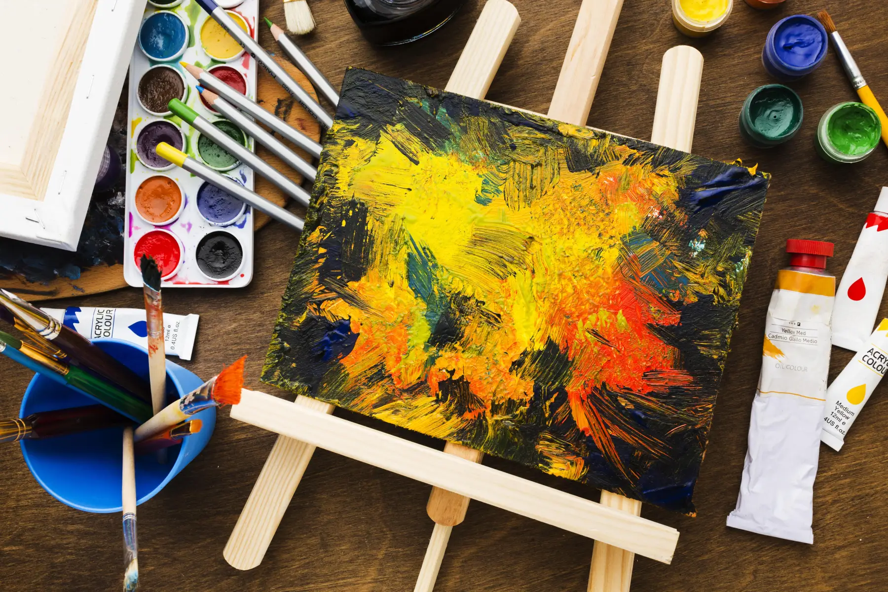 Vibrant abstract acrylic painting on an easel surrounded by artist paints, brushes and colour pencils